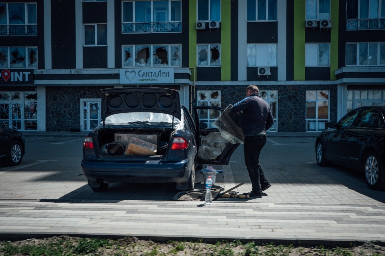 A photo of a man repairing his car in the middle of a parking lot in front of some shops.