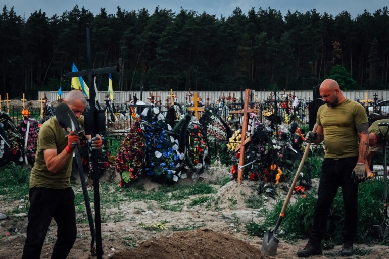 A photo of two men in a graveyard, the one on the right is holding two spades and the one on the right is holding a spade in the ground.