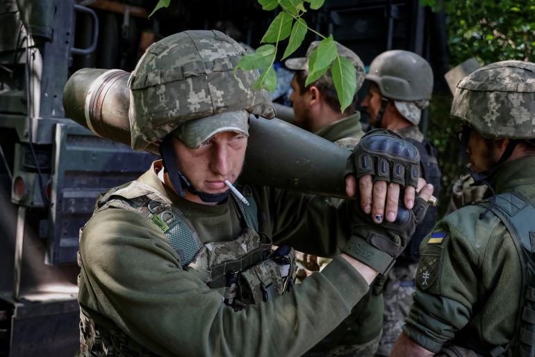 A Ukrainian service member holding a shell for an M777 Howitzer on his shoulder with other Ukrainian service members preparing shells for a M777 Howitzer in the background.