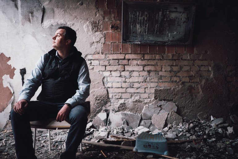 A photo of a man sitting in a room with crumbling walls and floors.