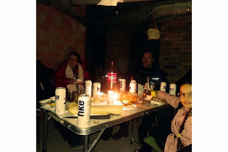 A photo of a group of people sitting around a table with food and drinks in dark basement with a light in the middle of the table.