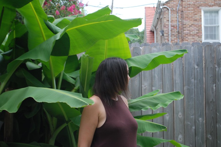 A photo of a woman standing next to a large plant outdoors in front of a fence to a house.