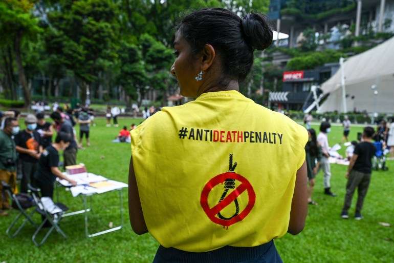 A woman with her back to the camera in a yellow T-shirt reading anti-death penalty and showing a red line through a noose