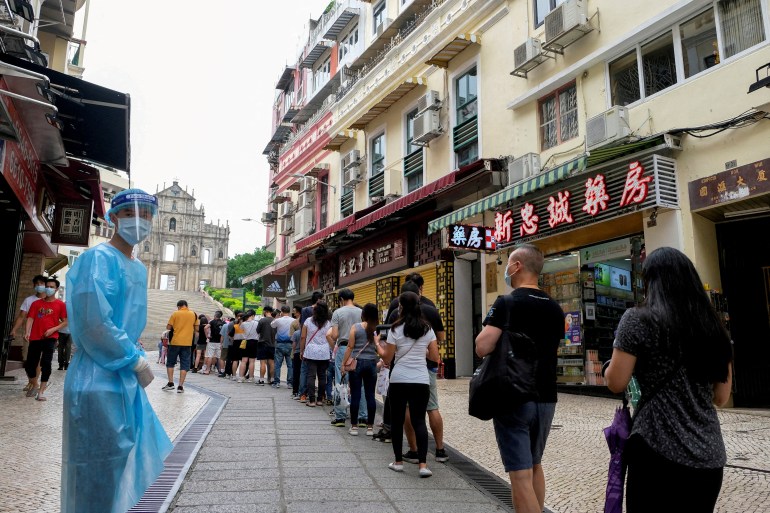People queue up on a street in Macau for COVID testing watched over by someone dressed in blue personal protective equipment.