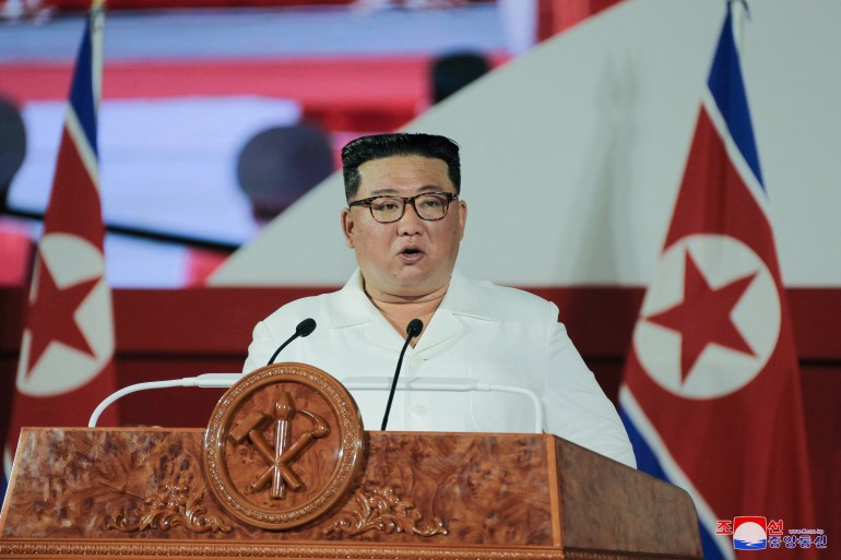 Kim Jong Un in a white open-necked shirt delivering his speech at a lectern with a North Korean flag behind him.