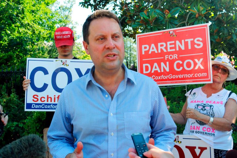 Dan Cox talks to reporters with some supporters holding his campaign signs in the background.
