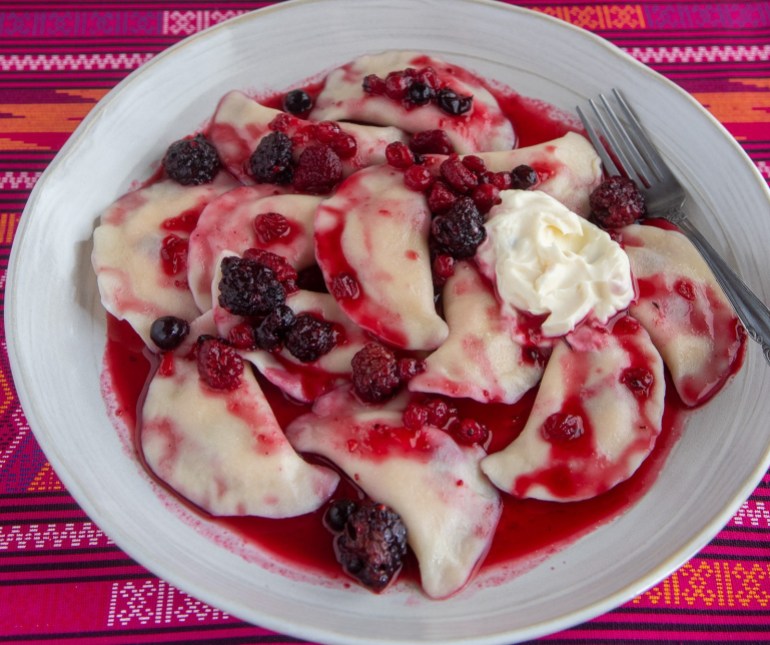 A photo of a plate of sweet dumplings with berry compote poured over top and a dollop of sour cream