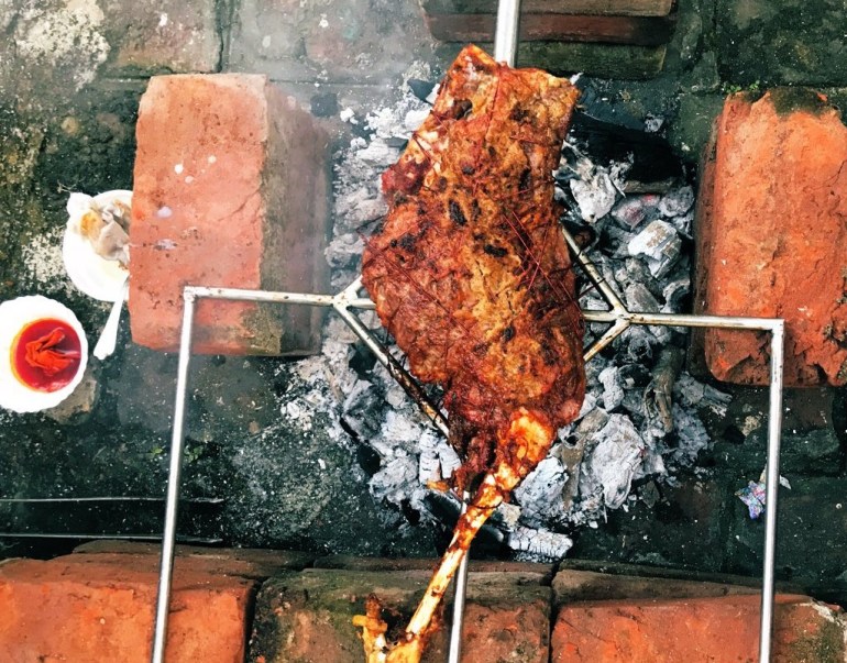 A leg of lamb on a makeshift brick barebecue, with two bowls of basting pastes on the ground near it