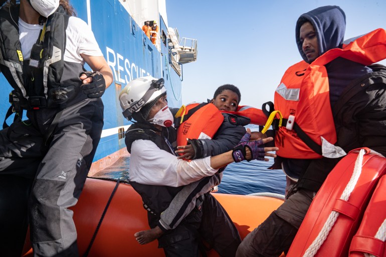  Mediterranean Sea near to Libya, 23 April 2022, cultural mediator, crew member and translator, Nejma Banks, holds a child who has been rescued as she coordinates the disembarkation of survivors from her boat. Each survivor moves to the bow of the rescue boat, where they must climb a ladder onto the ship. The MSF crew hold them at all times to ensure that nobody slips in the water. Credit: Lexie Harrison-Cripps