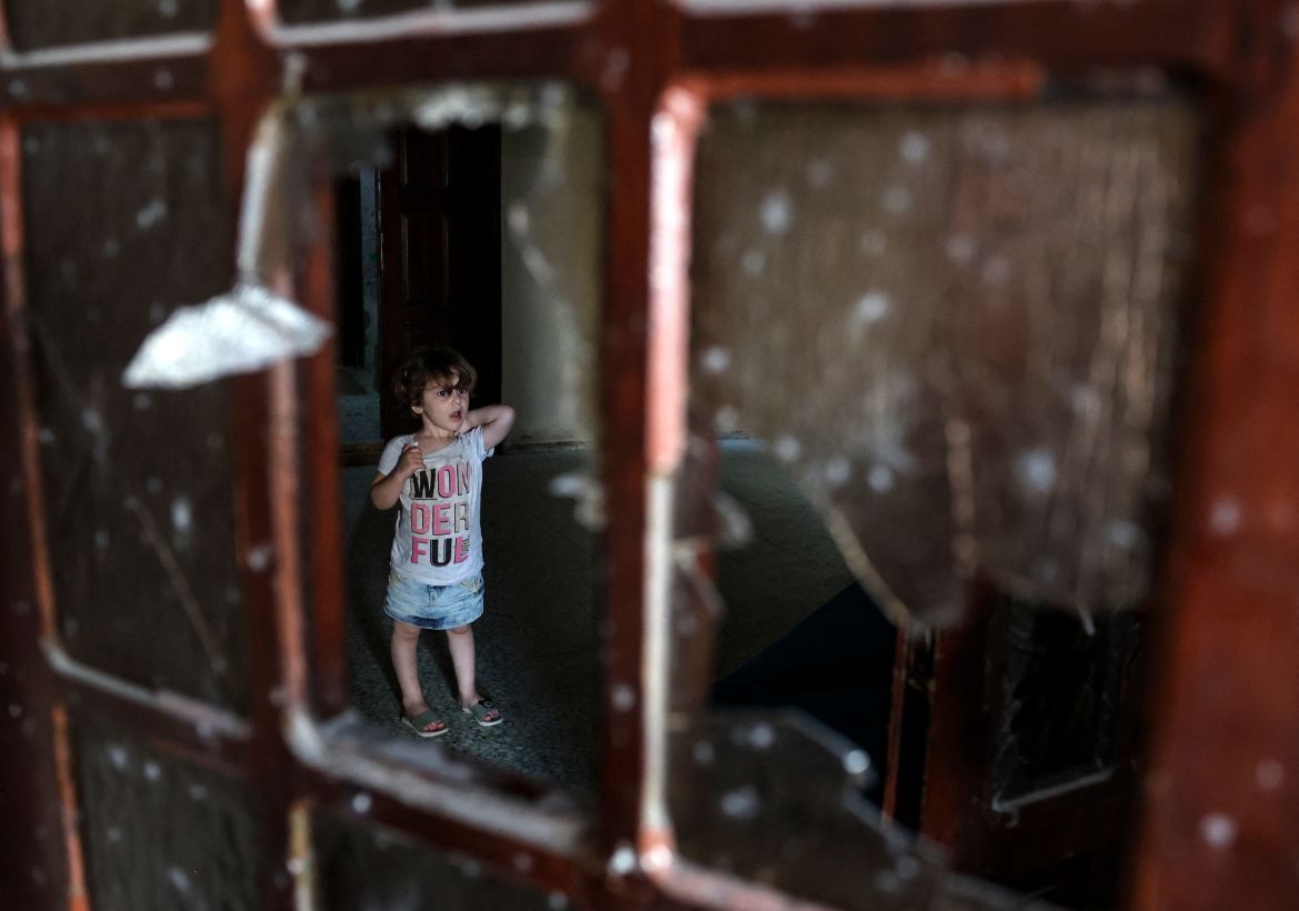 A Palestinian child stands behind a shattered window inside a building damaged