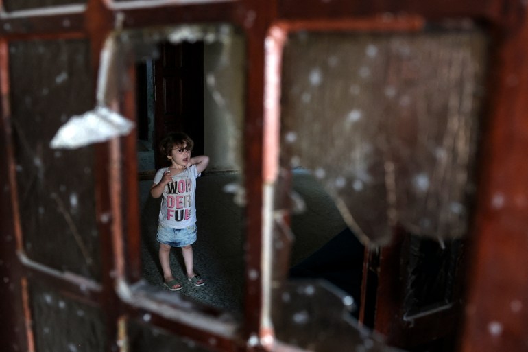 A Palestinian child stands behind a shattered window inside a building damaged