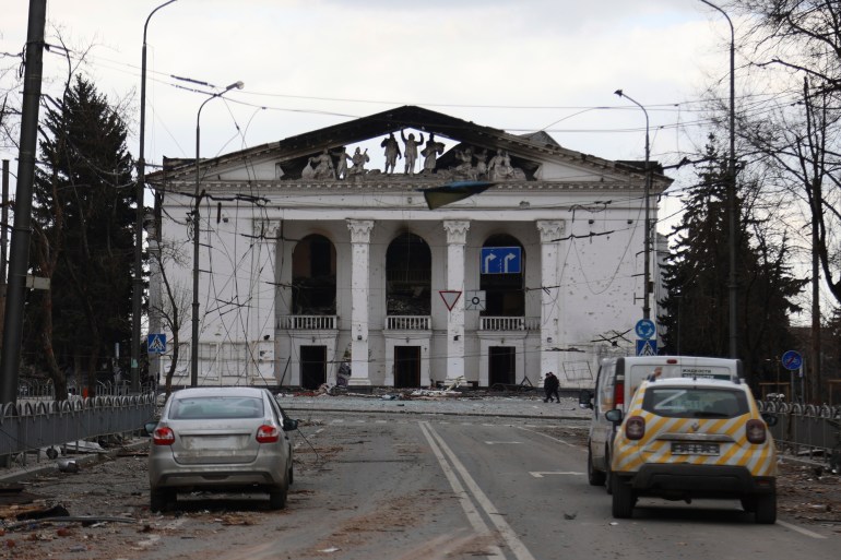 A photo of the outside of the damaged Donetsk Academic Regional Drama Theatre building with three cars in front of it.