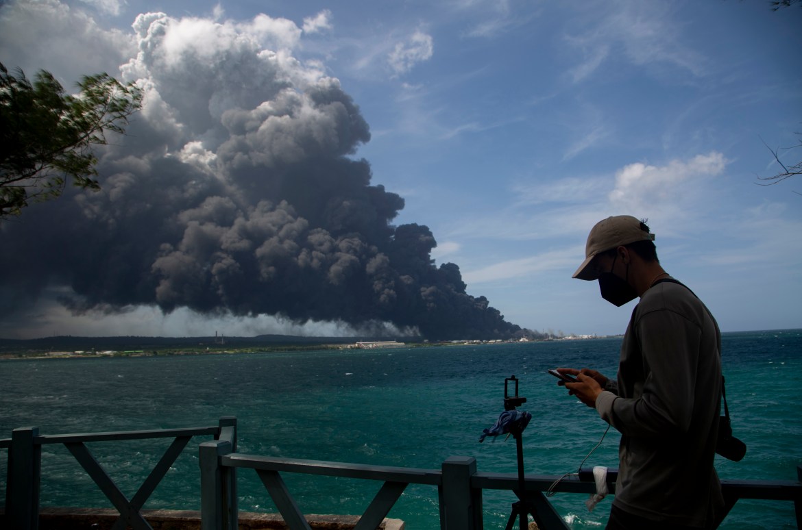 A man types on his cellphone near a huge plume of smoke rising from the Matanzas Supertanker Base as firefighters work to quell the blaze which began during a thunderstorm in Matanzas, Cuba
