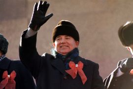 Gorbachev in black hat, coat and gloves with a red ribbon on his lapel waves from the parade review stand of the Lenin Mausoleum in November 1987