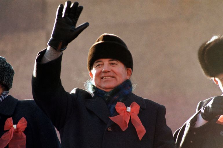 Gorbachev in black hat, coat and gloves with a red ribbon on his lapel waves from the parade review stand of the Lenin Mausoleum in November 1987