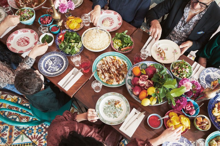 Overhead view of a family sharing an Afghan meal at a long table with bowls of fruit and herbs, platters of rice and kebab