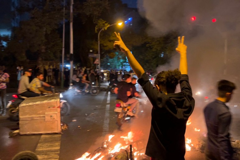 Woman in Iran raises her arms and displays the victory sign as protesters mill around her and items burn on the ground