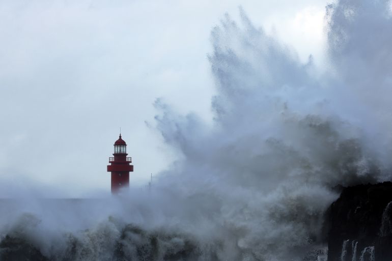 A giant wave crashes against the coast of Jeju island with the top of a lighthouse visible behind