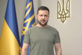 Zelenskyy in dark green shirt with Ukrainian flags behind him