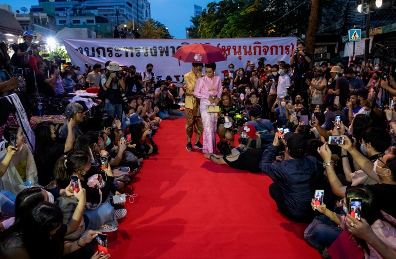 A pro-democracy protester in a pink silk dress, accompanied by a friend holding an umbrella over the protester's head, walks a mock red carpet at a Bangkok protest.