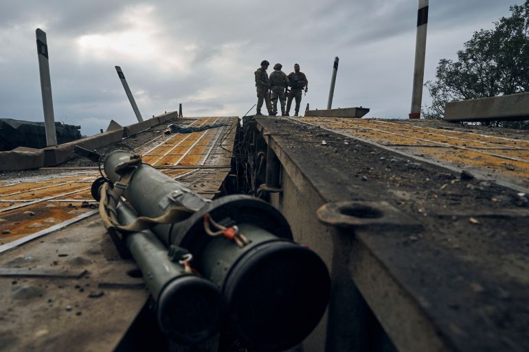 Three Ukrainian soldiers stand against the sky at the top of a destroyed bridge in Izyum with military equipment lying in the foreground