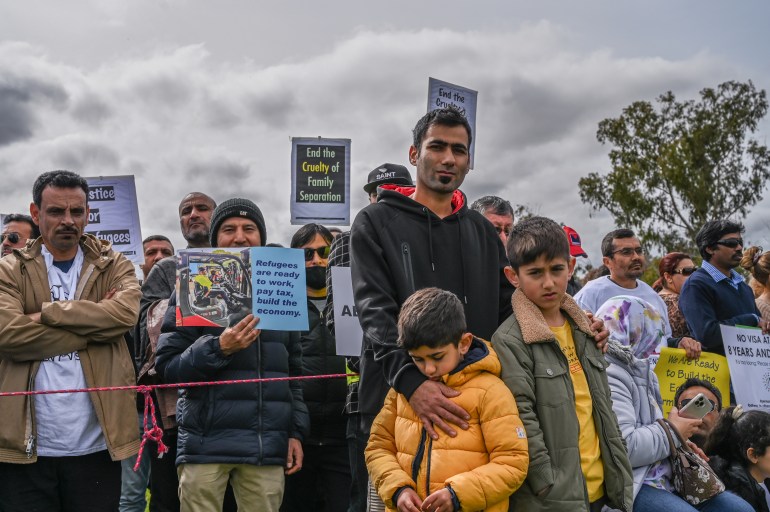 Un tată și cei doi băieți ai săi, unul privind cu tristețe la aparatul de fotografiat și cel mai mic privind în pământ.  participa la un protest pentru vizele de protecție permanentă în Canberra.  Alți protestatari cu bannere stau în spatele lor.