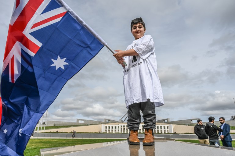 Un tânăr refugiat care poartă un tricou alb cu cuvântul „Viză permanentă” în pantaloni negri și gri, arborează un steag australian în fața Parlamentului din Canberra