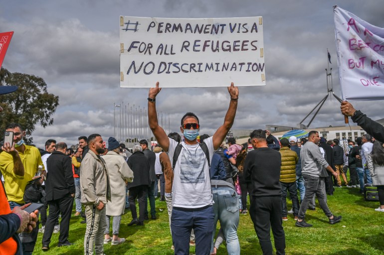 Un bărbat într-un tricou alb și pantaloni albaștri stă cu protestatarii ținând în sus o pancartă pe care scrie: #Viză permanentă pentru toți refugiații.  Fără discriminare