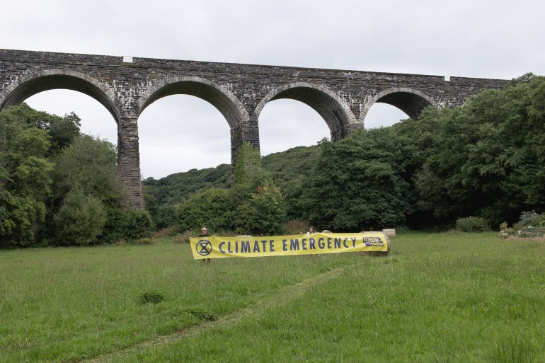A photo of two people in the distance holding a large poster/banner with the words "climate emergency" on it with a bridge behind them.