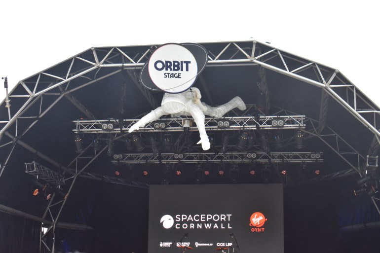 A photo of a stage with a large banner/poster with the words "spaceport Cornwall" on it and a figure of someone wearing a spacesuit floating above the stage lights.