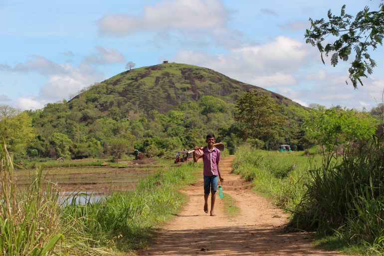 A young farmer walks smiling down a dirt path towards the photographer, with a hill in the background set against a bright blue sky