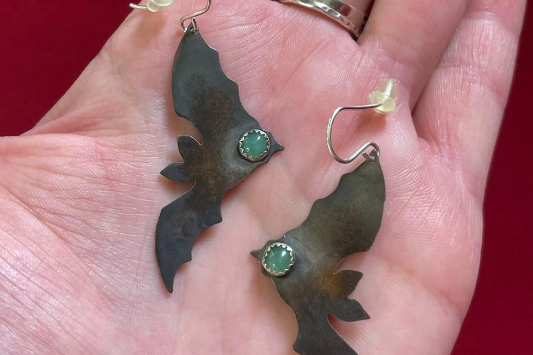 A photo of a person holding a pair of earrings with a bat shape cutout hanging off the earrings with a green stone on the end of the bat.