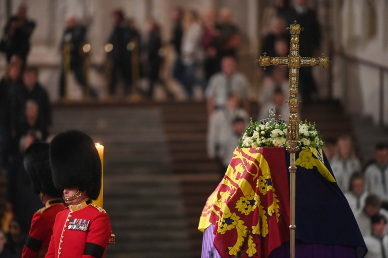 a soldier in red ceremonial dress and bearskin stands guard by the queen's coffin as people pay their final respects