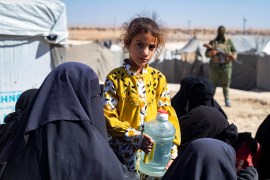 A girl looks on while holding a water container as members of the Syrian Kurdish Asayish security forces inspect tents at the Kurdish-run al-Hol camp