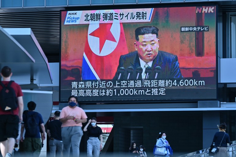 People walking on street past large screen with North Korean showing Kim Jung Un speaking.