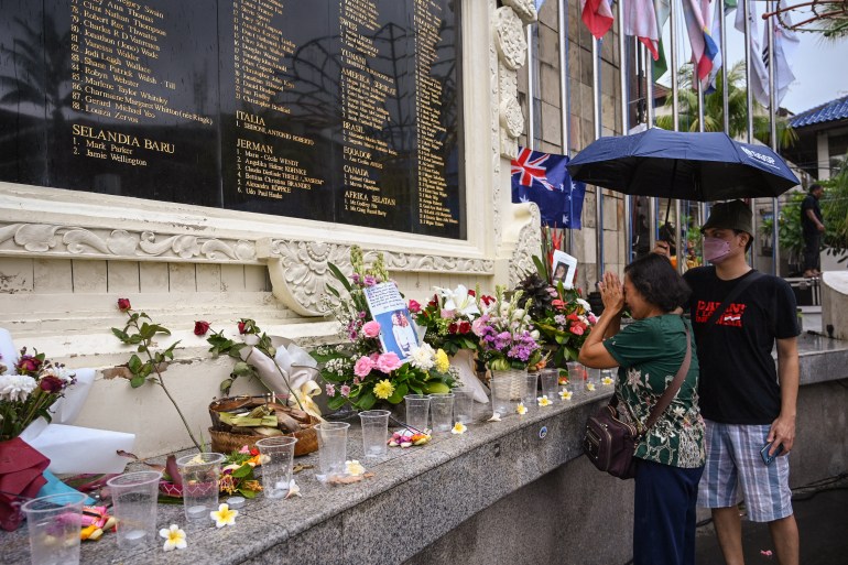 A woman prays after laying a flower at a memorial site, next to a line of other flowers. Man with umbrella walks by