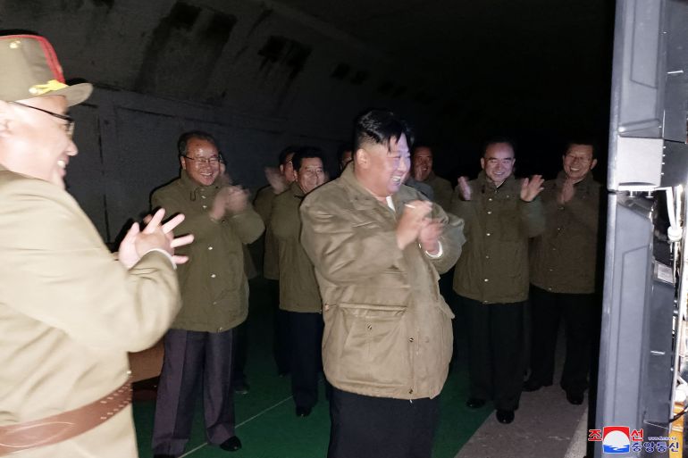 Kim Jong un in a brown jacket smiles and clasps his hands after watching the launch of two cruise missiles