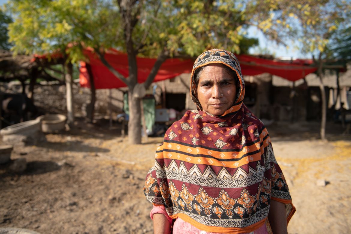 Kasim’s wife, Zeenat Bibi, stands in front of her damaged home. She and her family set up a temporary shelter using tarp and sticks taken from trees nearby, as their house is unsafe.