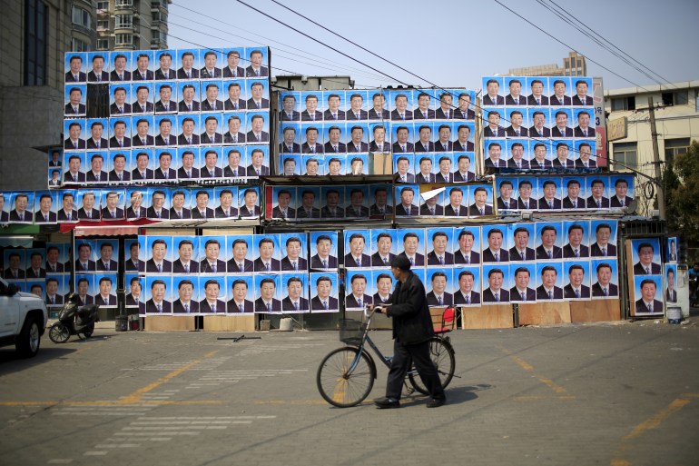 Man with bicycle walks past a high wall plastered over with about one hundred of the same posters of Xi Jinping's face