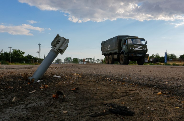 Российский военный грузовик с буквой Z на переднем плане направляется к неразорвавшемуся снаряду, похожему на дротик, застрявшему в земле.