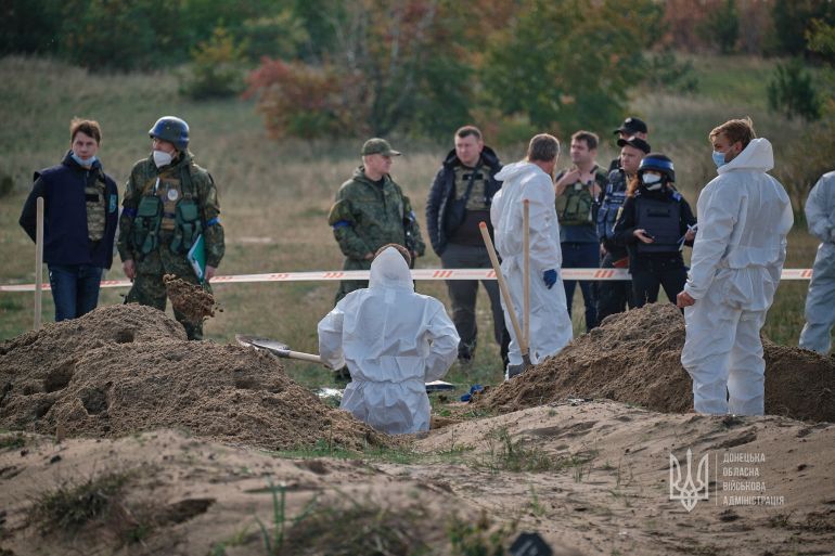 Forensic technicians in white and officers work, one standing in a hole, at what appears to be a mass grave declared by regional governor Pavlo Kyrylenko as found in the town of Lyman