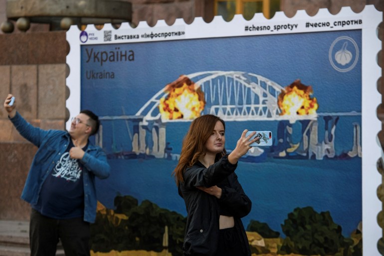 A man and a woman take selfies in front artwork depicting Kerch bridge on fire