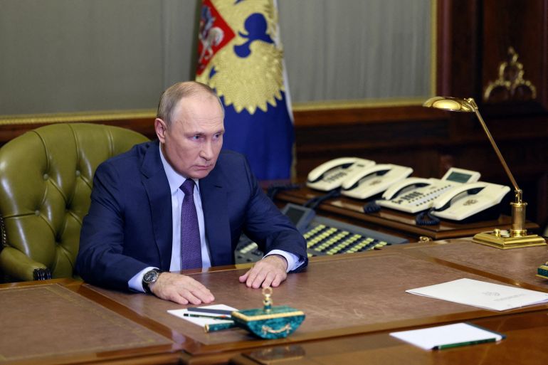 Vladimir Putin sitting at an almost empty wooden desk with a flag and some white phones behind him.