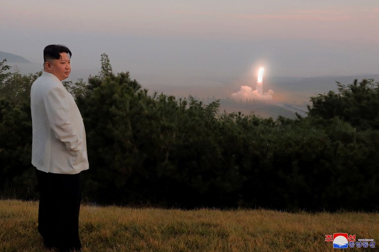 Kim Jong Un in white jacket stands on a hillside watching an explosion in the distance