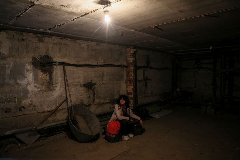 A woman shelters in an empty basement, except for a steel crate and shovel hanging on wall. She has a red backpack.