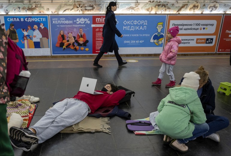 Person lying on rug on the ground, watching something on their laptop. Mother and child are squatting facing each other and doing something on the ground