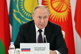 Vladimir Putin sitting behind podium with flags behind him