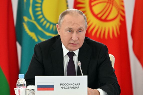 Vladimir Putin sitting behind podium with flags behind him