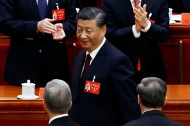 Xi smiling at two men, whose backs of heads appear in the photo, as he walks past wearing a red name tag with a yellow hammer and sickle on it.