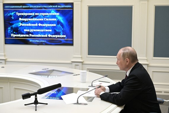 Putin sits at round table wearing black suit and looking at screen to his right.